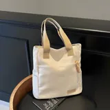 Cartera tipo shopper color beige con bolsillos laterales y un bolsillo frontal con tirador de cordón trenzado. Cuenta con asas superiores de lona.