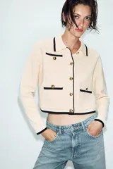 Chaqueta de punto color crema con ribetes en contraste negro, cuello solapa, manga larga y cierre frontal con botones dorados. Presenta bolsillos de plastrón en el delantero con detalle de botones.