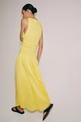Vestido largo amarillo de algodón, con cuello barco y mangas sisa. Presenta diseño drapeado en los laterales y falda de corte evasé con volumen en el bajo.
