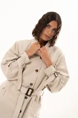 Trench corto color beige con cuello alto, cierre frontal de botones y cinturón ajustable en la cintura. Presenta mangas largas con detalles de tiras ajustables en los puños y bolsillos laterales.