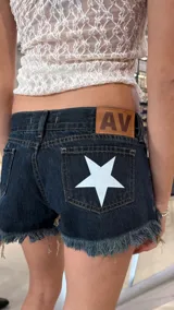 Short de jean azul oscuro con tiro bajo, caracterizado por un diseño de estrella blanca estampada en el bolsillo trasero y bordes deshilachados en el ruedo. Incluye una etiqueta de cuero sintético con las iniciales AV en la parte posterior.