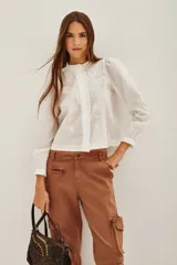 Camisa blanca de algodón con cuello alto, cierre frontal de botones ocultos y detalles de bordado floral en el pecho. Presenta mangas largas con puños elásticos y volados decorativos en la parte frontal.