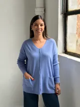 Sweater de punto color azul lavanda, con escote en V, mangas largas con puños acanalados y dos bolsillos frontales tipo parche. Presenta una costura central vertical visible.