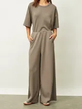 Mono de una pieza color gris topo, con cuello redondo, mangas cortas y piernas anchas. Presenta cintura elástica y bolsillos laterales.