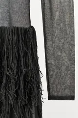 Vestido midi negro, semitransparente, de manga larga y cuello redondo. La parte superior presenta un tejido de punto fino con costuras que simulan un bustier. La falda está compuesta por capas de flecos largos y densos.