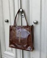 Cartera tipo tote de cuero marrón con acabado brillante, diseño con costuras artesanales en los bordes y detalle de trenzado decorativo en el bolsillo frontal. Incluye doble asa de hombro y logo grabado.
