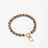 Accesorio decorativo para cartera compuesto por una cadena de cuentas de ojo de tigre intercaladas con pequeñas esferas doradas, finalizado con un mosquetón metálico dorado.