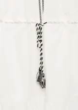 Vestido corto de algodón en color blanco, con diseño evasé y detalles bordados en contraste negro en hombros, mangas y cuello. Presenta mangas largas con puños elásticos, cuello redondo con abertura frontal y cintura elástica ajustable mediante un lazo con borlas.