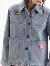 Chaqueta de denim de algodón color azul con efecto lavado, corte recto y cierre frontal con botones. Presenta bolsillos aplicados en el frente y bordados de una abeja y flores.
