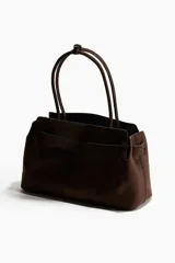 Bolso tote de acabado suave, color marrón oscuro, con asas tubulares y detalle de correa frontal con hebilla metálica dorada.