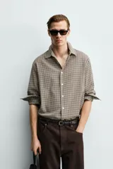 Camisa de corte relajado confeccionada en tejido de algodón con textura irregular y estampado de cuadros pequeños en tonos tierra. Presenta cuello solapa, cierre frontal de botones y mangas largas con puños abotonados.