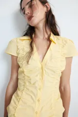 Top de cuello solapa y escote en pico, con manga corta y cierre frontal con botones. Presenta un diseño ajustado con detalles de frunces en el delantero.