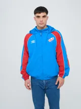 Campera deportiva Umbro del Club Nacional de Football, modelo Venti, para hombre. Diseño en color blanco con mangas azules y detalles de líneas rojas. Cuenta con capucha con cordón ajustable, cierre frontal y bolsillos. Presenta el logo de Umbro y el escudo del club bordados en el pecho.