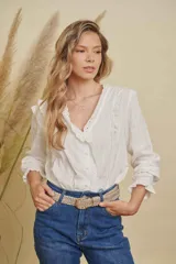 Camisa blanca de manga larga con diseño de volados en los hombros y cuello, confeccionada en tela con textura de rayas verticales y detalles bordados en los bordes. Presenta cierre frontal con botones y puños elásticos con volados.