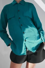 Camisa de corte fluido con cuello solapa y manga larga. Presenta cierre frontal con botones y dos bolsillos de plastrón con solapa en el pecho. El diseño cuenta con bajo redondeado y costuras verticales decorativas.
