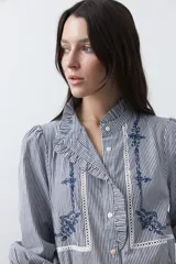 Camisa de rayas verticales con cuello mao, detalle de volados en el escote, bordados florales y recortes con calados. Posee mangas largas abullonadas con puños ajustados y cierre frontal con botones.