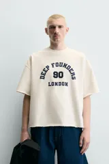 Sudadera boxy fit de color beige con interior de rizo, cuello redondo y manga corta. Presenta un estampado contrastante en el frente con la inscripción "Deep Founders London 90" y acabados irregulares en los bordes.