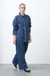 Sobrecamisa de jean negra de silueta relajada, con tachas aplicadas en líneas orgánicas que recorren la prenda. Tiene cuello camisero, cierre frontal con botones a presión y mangas largas.
