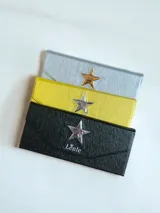 Estuche para lentes plegable con diseño rígido y cierre magnético. Presenta una estrella metálica decorativa en la solapa frontal y el logo de la marca estampado. El interior está forrado en material suave para proteger los cristales.