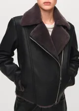 Chaqueta estilo aviador de color negro con interior de piel sintética gris oscuro, cierre frontal cruzado con cremallera y solapas grandes.