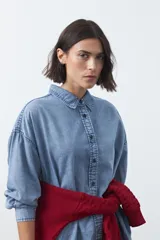 Camisa de jean en tencel con silueta amplia, hombros caídos, cuello clásico y cierre frontal con botones. Presenta ruedo curvo con tajos laterales y pliegue en la espalda.