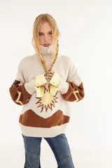 Sweater de punto en color beige con diseño de intarsia que representa un sol y montañas en tonos marrón y amarillo. Presenta cuello alto con cierre frontal, mangas largas y corte holgado.