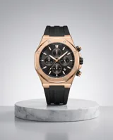 Reloj cronógrafo con caja de aleación metálica en tono oro rosa y esfera negra. Presenta indicadores de hora, subesferas funcionales y correa de caucho negra con cierre de hebilla.