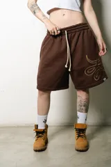 Bermuda estilo baggy de algodón color marrón oscuro, con cintura elástica y cordón blanco. Presenta un gran logo estampado en tono gris claro en la pierna derecha.