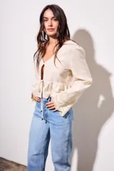 Blusa beige de viscosa y lino, con escote en V, mangas largas acampanadas con detalles bordados tipo broderie, y cierre frontal con lazo para anudar.