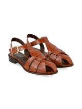 Sandalias planas de cuero color marrón coñac, estilo huarache con tiras entrelazadas en la parte delantera, tira en T y cierre de hebilla metálica dorada en el tobillo. Cuenta con un tacón bajo cuadrado.