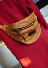 Riñonera de cuero vacuno color camel, con cierre principal y tirador de cuero, correa regulable y parche con logo grabado en el frente.