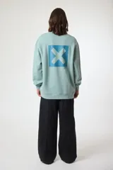 Buzo de cuello redondo en color crema, con diseño de logo cuadrado en el pecho que incluye una X blanca sobre fondo azul y caracteres japoneses. Presenta puños y ruedo acanalados.