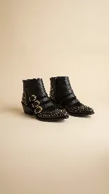 Botas cortas de cuero negro con diseño estilo western, decoradas con múltiples tachas metálicas en tono dorado y plateado. Presentan tres correas horizontales con hebillas doradas sobre el empeine y cierre lateral.