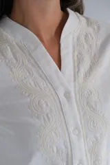 Blusa blanca de manga larga con cuello mao y cierre frontal de botones. Presenta detalles de bordados decorativos en los laterales del frente y puños anchos.