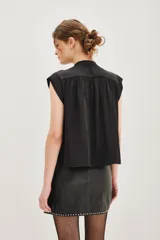 Camisa sin mangas de satén negro, con cuello mao y cierre frontal oculto con un botón decorativo en forma de corazón en la parte superior. Presenta un corte recto y fluido.