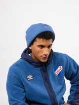 Gorro tipo beanie de tejido de punto color blanco crema, con parche frontal que incluye el logo de Umbro y la inscripción Nacional.