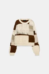 Jersey de punto con cuello redondo y manga larga, caracterizado por un diseño de parches en tonos tierra y crema. Presenta una textura variada con detalles de flecos y tejido en relieve.