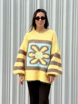 Buzo de tejido de punto con textura peluda, de corte holgado y mangas amplias. Presenta un diseño geométrico con una flor grande estampada en el centro, combinando tonos fucsia, rosa y marrón en un patrón de bloques y rayas.