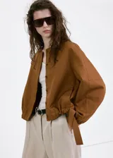 Chaqueta de diseño corto en tono tostado, confeccionada con mezcla de lyocell, algodón y lino. Presenta cuello redondo, mangas largas abullonadas, elásticos en los laterales y cierre frontal de botonadura visible.