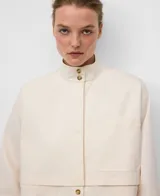 Camisa de mujer color crudo, de corte recto, con cuello chimenea abotonado y manga larga con abertura. Presenta botonadura frontal y bolsillos de plastrón con logo bordado en el pecho.