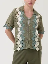 Sweater verde de tejido tipo crochet, con cuello camisero, mangas cortas y cierre frontal de botones. Presenta un diseño de bandas laterales con aplicaciones florales en tonos blancos y celestes.