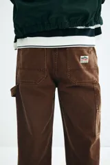 Pantalón worker marrón de sarga de algodón gruesa, con piernas rectas y corte relaxed fit. Presenta cintura clásica con cierre de botón, bolsillos al bies, bolsillo monedero, bolsillos traseros, trabilla funcional en un lateral y bolsillo cargo dividido en dos en el otro.