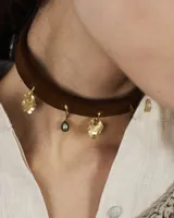 Gargantilla tipo choker de gamuza marrón con dijes colgantes de bronce o alpaca de diseño irregular y una perla natural de río. Posee un sistema de ajuste mediante tiras para atar en la parte posterior.