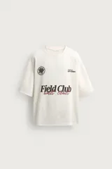 Remera de algodón color crudo, con textura de tejido abierto, cuello redondo y mangas cortas. Presenta estampas en contraste en el frente con los textos 'Field Club', 'West Coast', gráficos deportivos y un escudo circular.