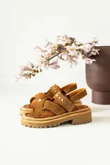 Sandalias de gamuza color tostado con diseño de tiras cruzadas en el empeine y tira trasera con cierre de hebilla. Presentan detalles de tachas metálicas circulares en las tiras y una suela de plataforma dentada de goma.