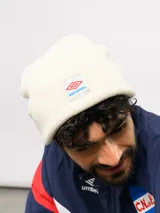 Gorro tipo beanie de tejido de punto color blanco crema, con parche frontal que incluye el logo de Umbro y la inscripción Nacional.