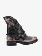 Bota corta de cuero bovino con acabado desgastado en tonos gris y negro. Presenta un diseño rebelde con múltiples correas ajustables con hebillas y tachas metálicas decorativas en la caña. Tiene un taco bajo y suela gruesa.