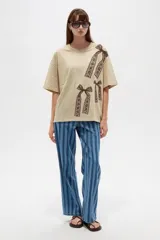 Remera oversize de algodón color beige con cuello redondo y mangas cortas. Presenta un estampado frontal con diseño de moños marrones estilo encaje.