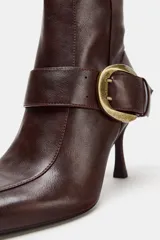 Bota de caña alta en color marrón oscuro, confeccionada en cuero con acabado liso. Presenta un diseño de punta fina, tacón alto fino y una tira decorativa en el tobillo con una hebilla metálica dorada. Incluye cierre lateral mediante cremallera.