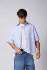 Camisa de manga corta a rayas celestes y blancas, de corte boxy, con cierre frontal de botones y pequeño estampado en el pecho.
